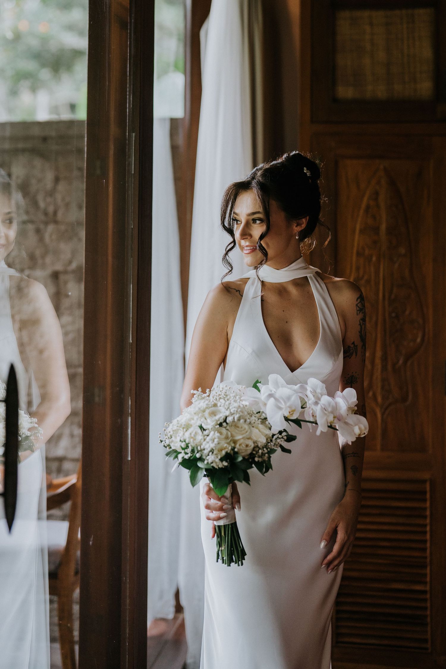 Bride in elegant white halter dress holds bouquet of white flowers by wooden window.