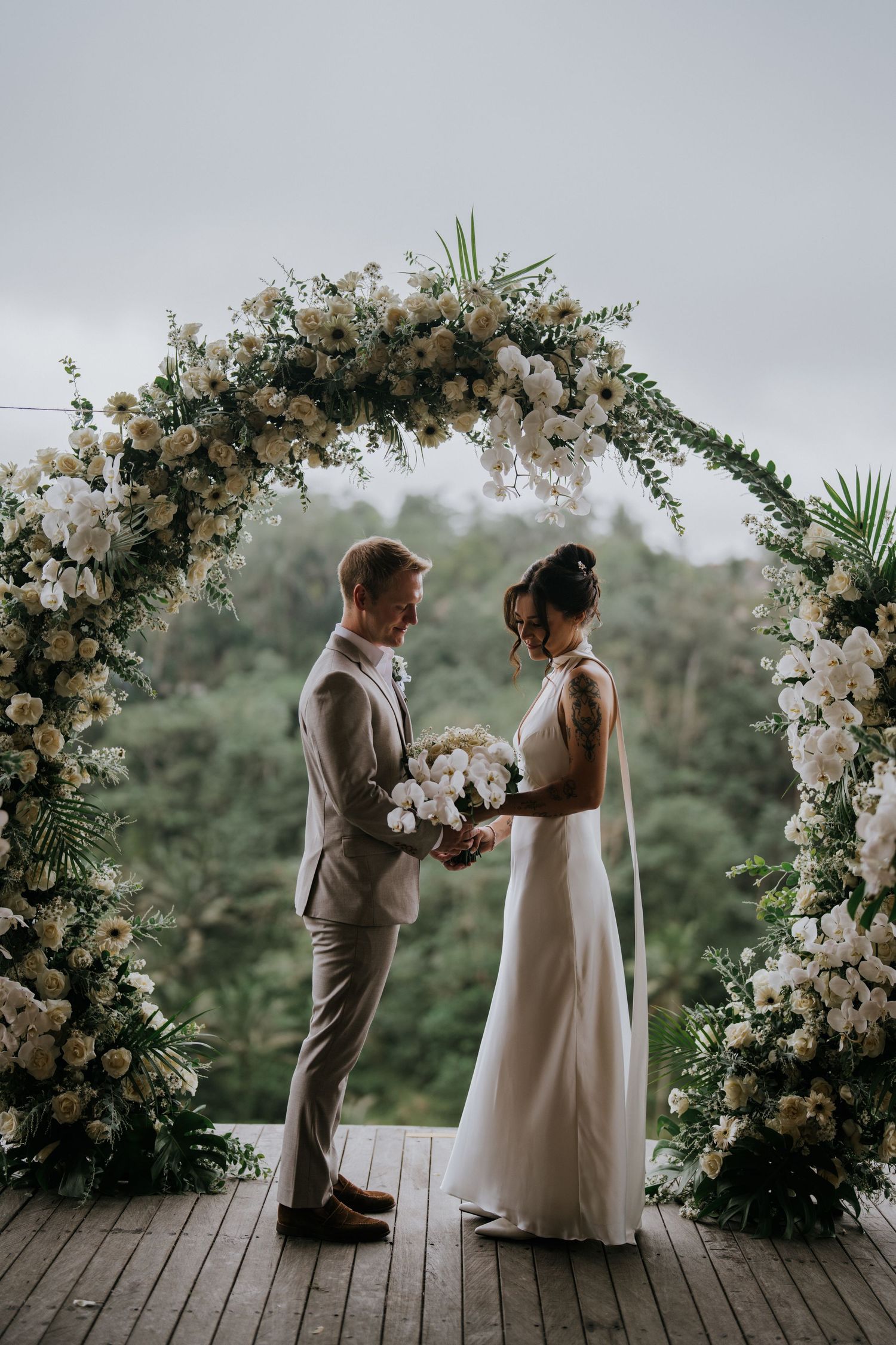 Bride in elegant white halter dress holds bouquet of white flowers by wooden window.