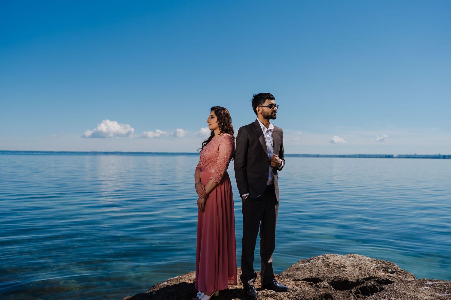 A couple stands on rocky coastal overlook admiring vibrant blue ocean views and horizon on a sunny day.