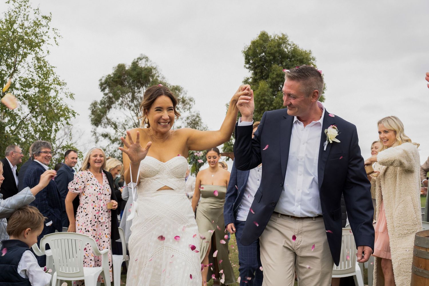 A happy couple celebrates with raised hands while walking down the aisle as wedding guests toss rose petals.
