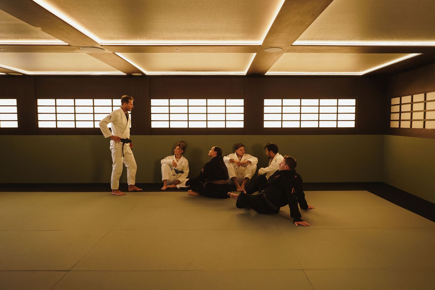 People practicing martial arts in a dimly lit dojo with wooden floors and traditional Japanese-style windows.