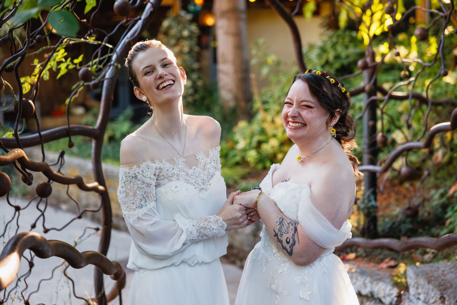 Two brides in white lace gowns laugh and hold hands outdoors in a sunlit garden setting.