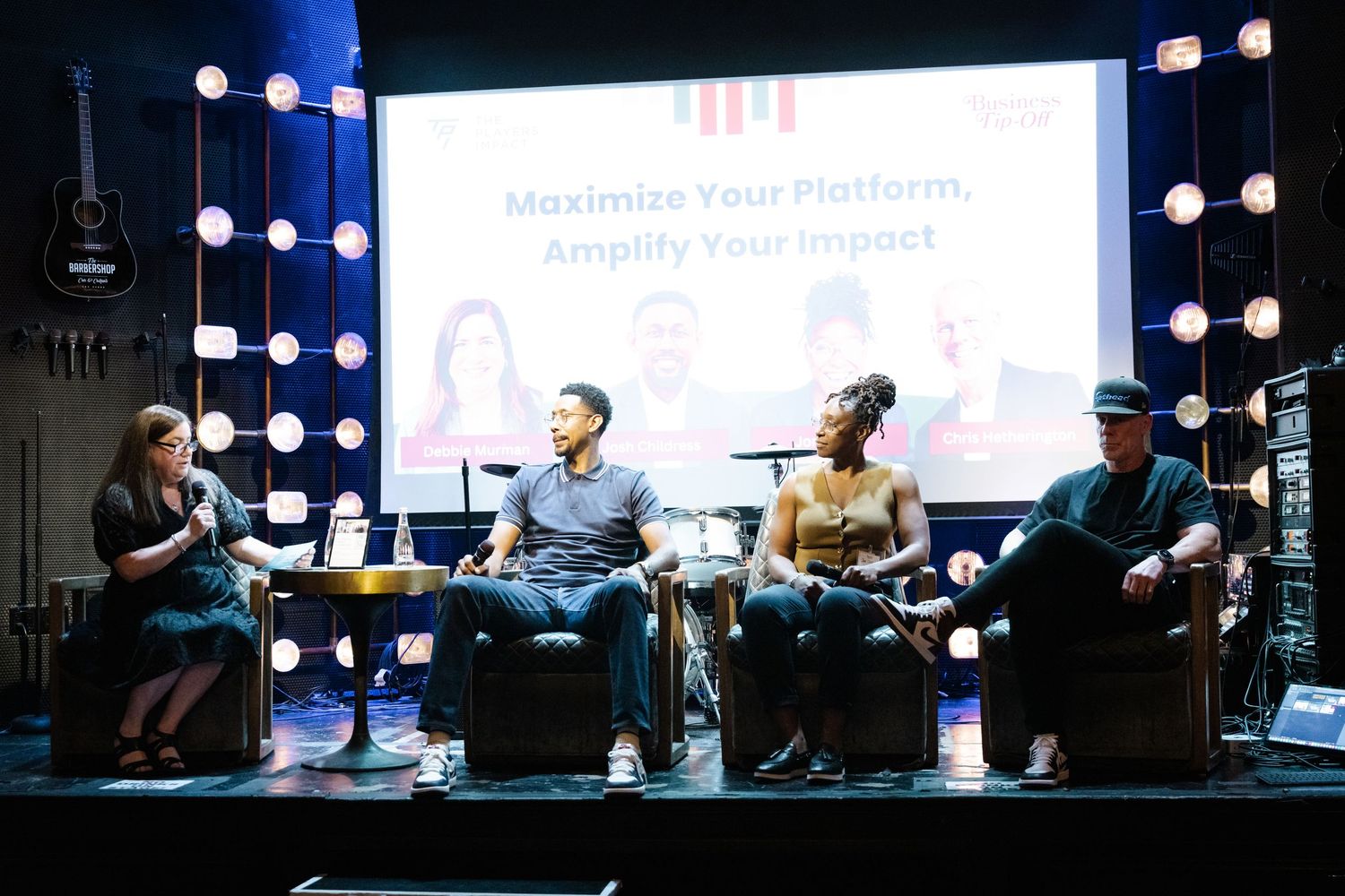 Panel discussion taking place on stage with four speakers seated in chairs under blue lighting.