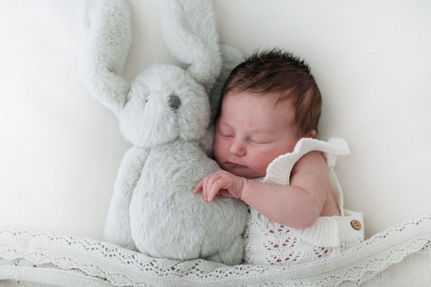 A sleeping newborn baby swaddled in white lace and surrounded by plush white teddy bears in a soft studio setting.