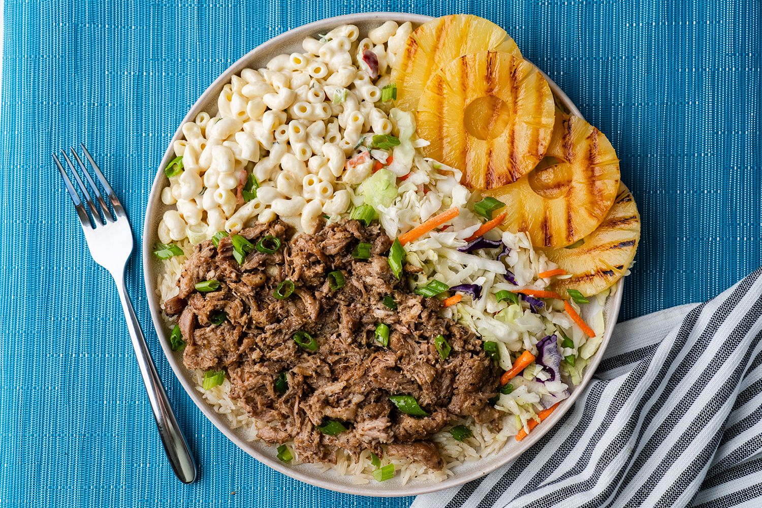 Kalua Pig Pulled pork, Hawaiian macaroni salad, coleslaw and grilled pineapple rings on a white plate against blue background.