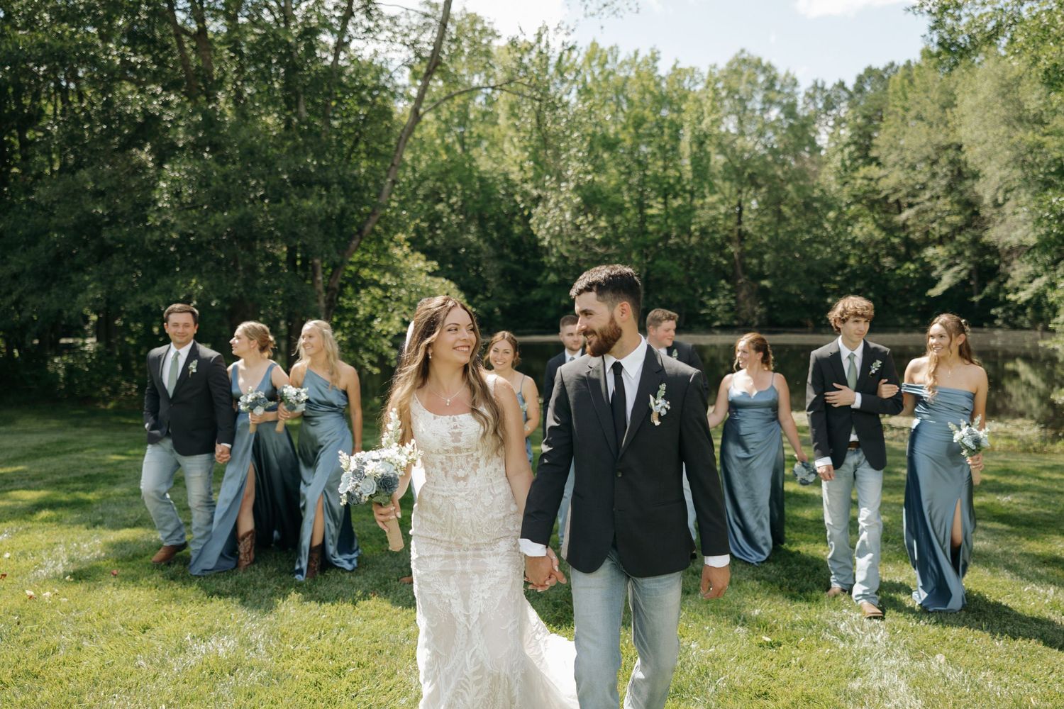 Wedding party in light blue attire stands on sunny lawn while newlyweds walk in foreground.