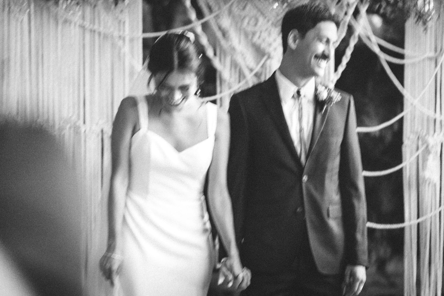A couple in formal wedding attire walk hand in hand through decorative white streamers in black and white.