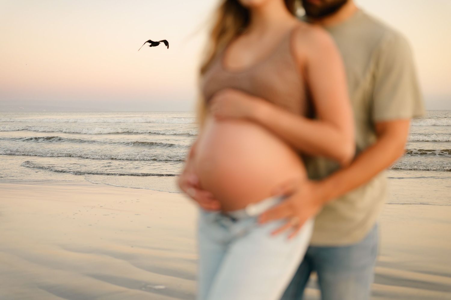 couple stand together with husband holding wife's pregnant bare belly in front of ocean waves, by Ventura and Ojai photographer