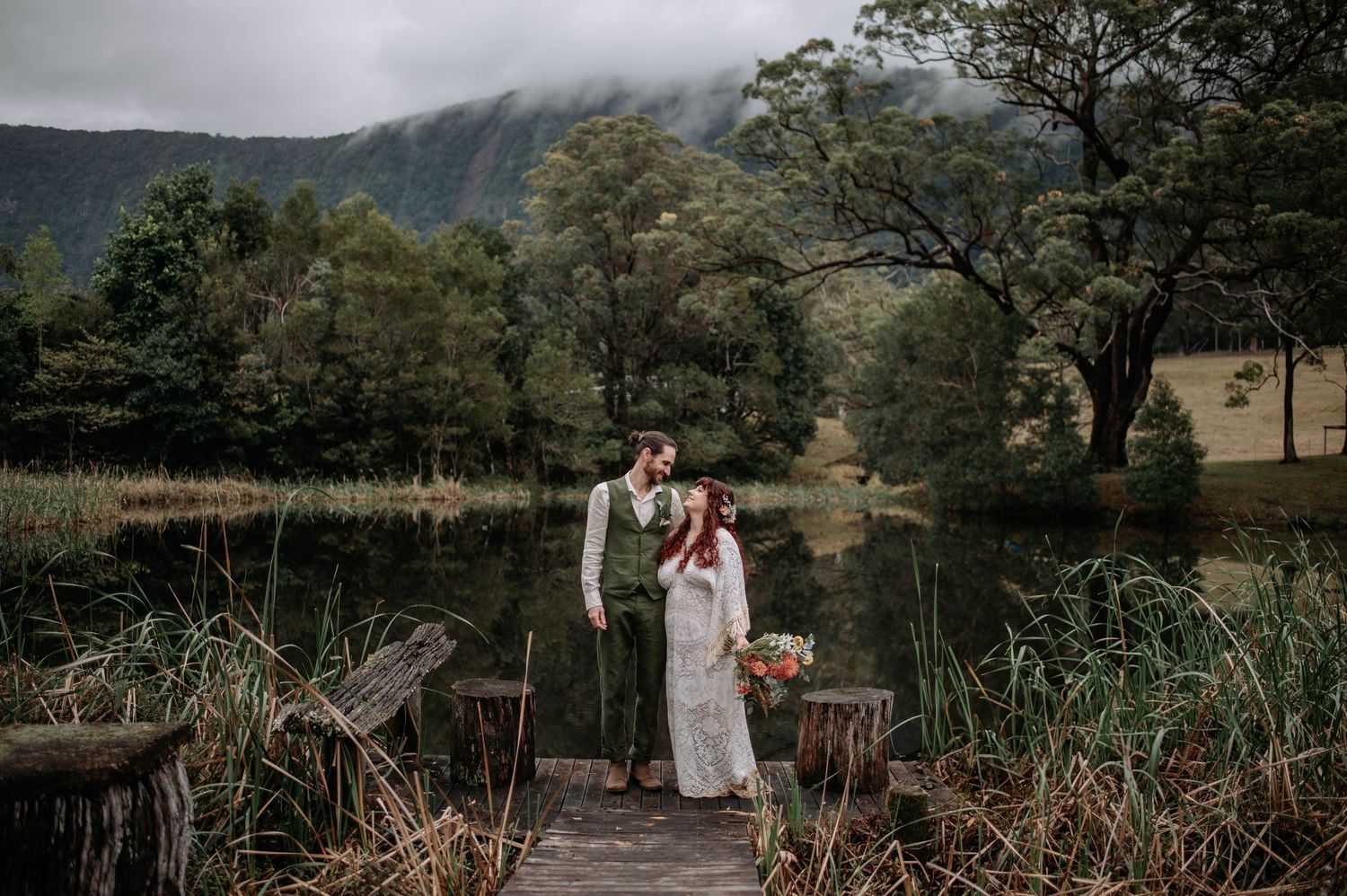 A wedding couple stands on a wooden bridge surrounded by tall grasses and mountains in a moody natural setting.