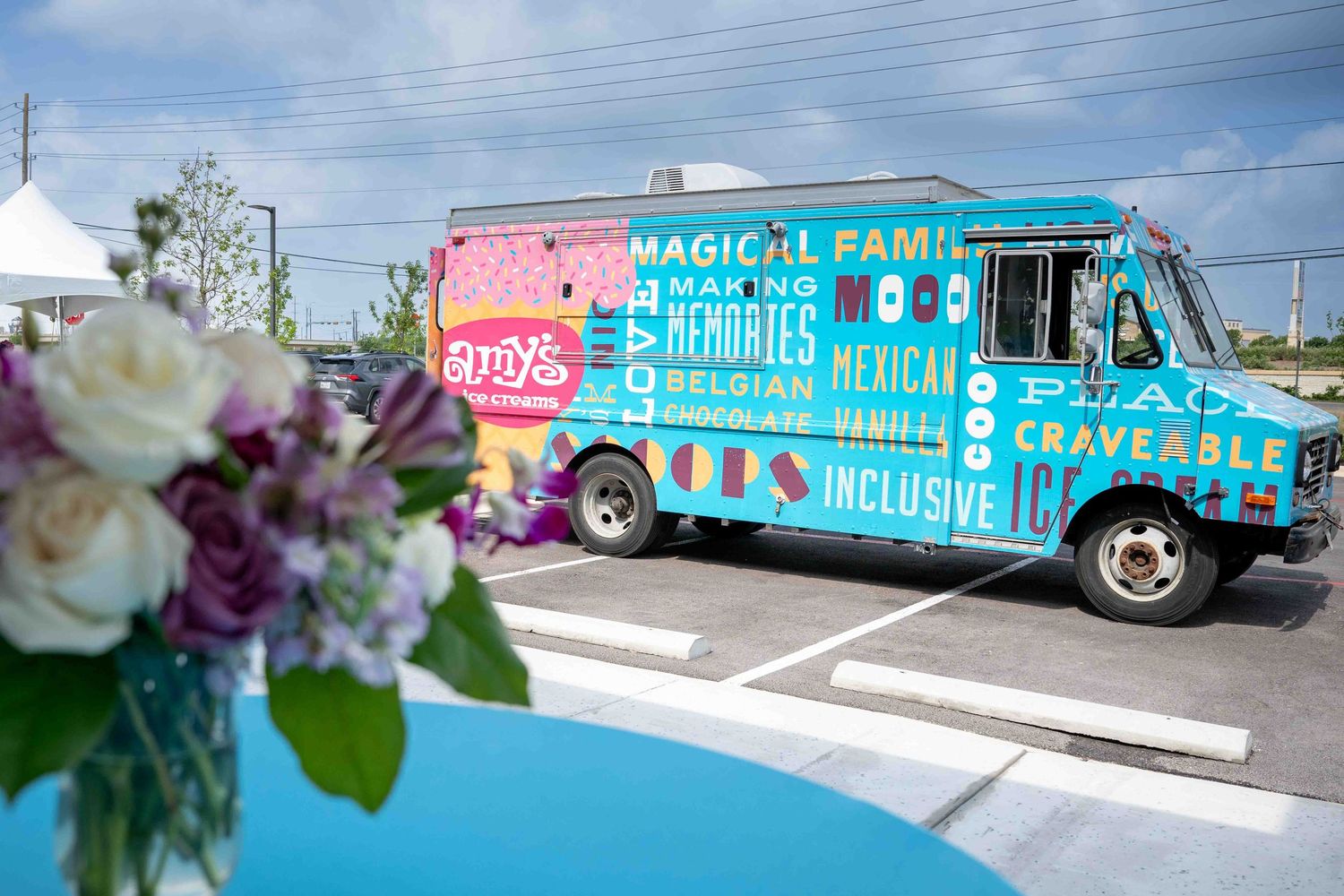 Bright turquoise food truck parked with floral centerpiece in foreground featuring purple and white flowers in a vase.
