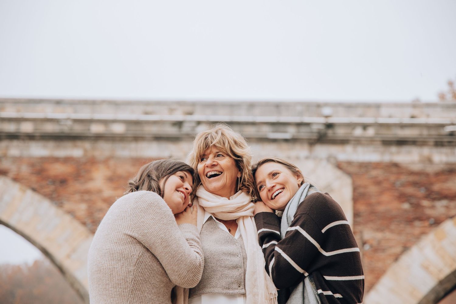 Three happy women laughing together outdoors near a stone bridge arch.
