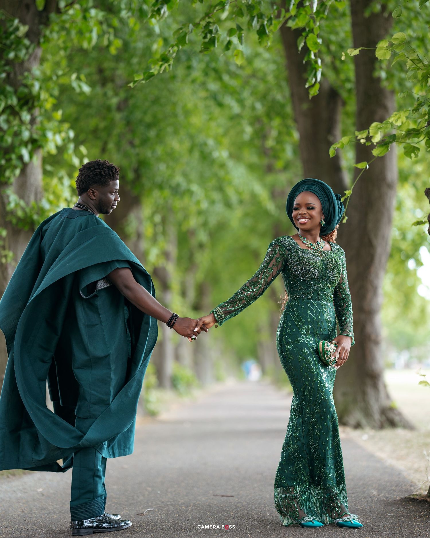 Couple in matching emerald green traditional Nigerian attire holding hands on a tree-lined pathway.