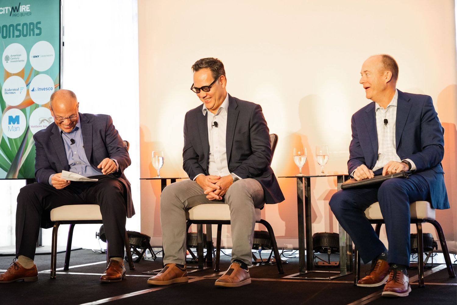 Three businessmen laughing and conversing on stage at a corporate conference event with sponsor banners.
