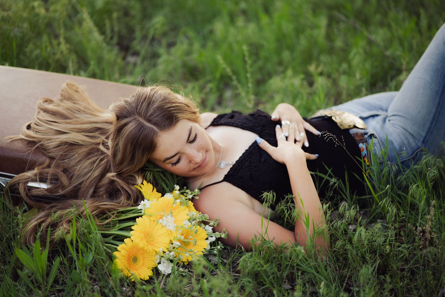 Young woman with long hair lies in green grass holding yellow gerbera daisy bouquet.
