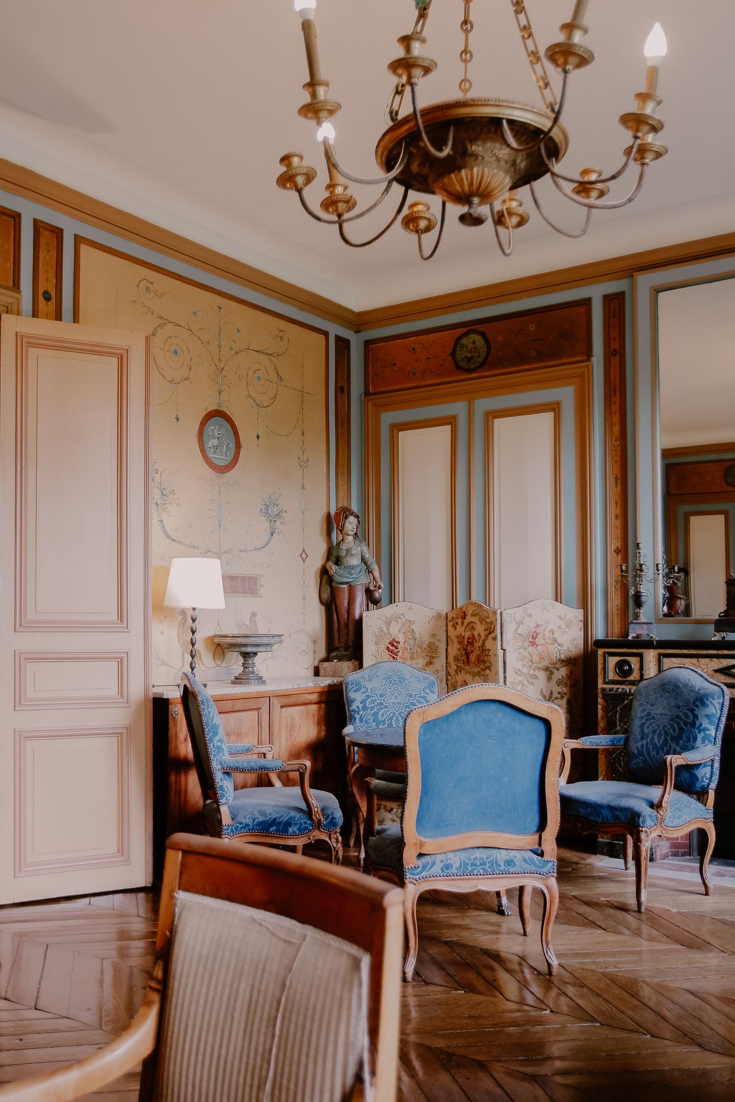 Elegant French salon with blue velvet Louis XV chairs, ornate wood paneling, chandelier, and decorative figurine.