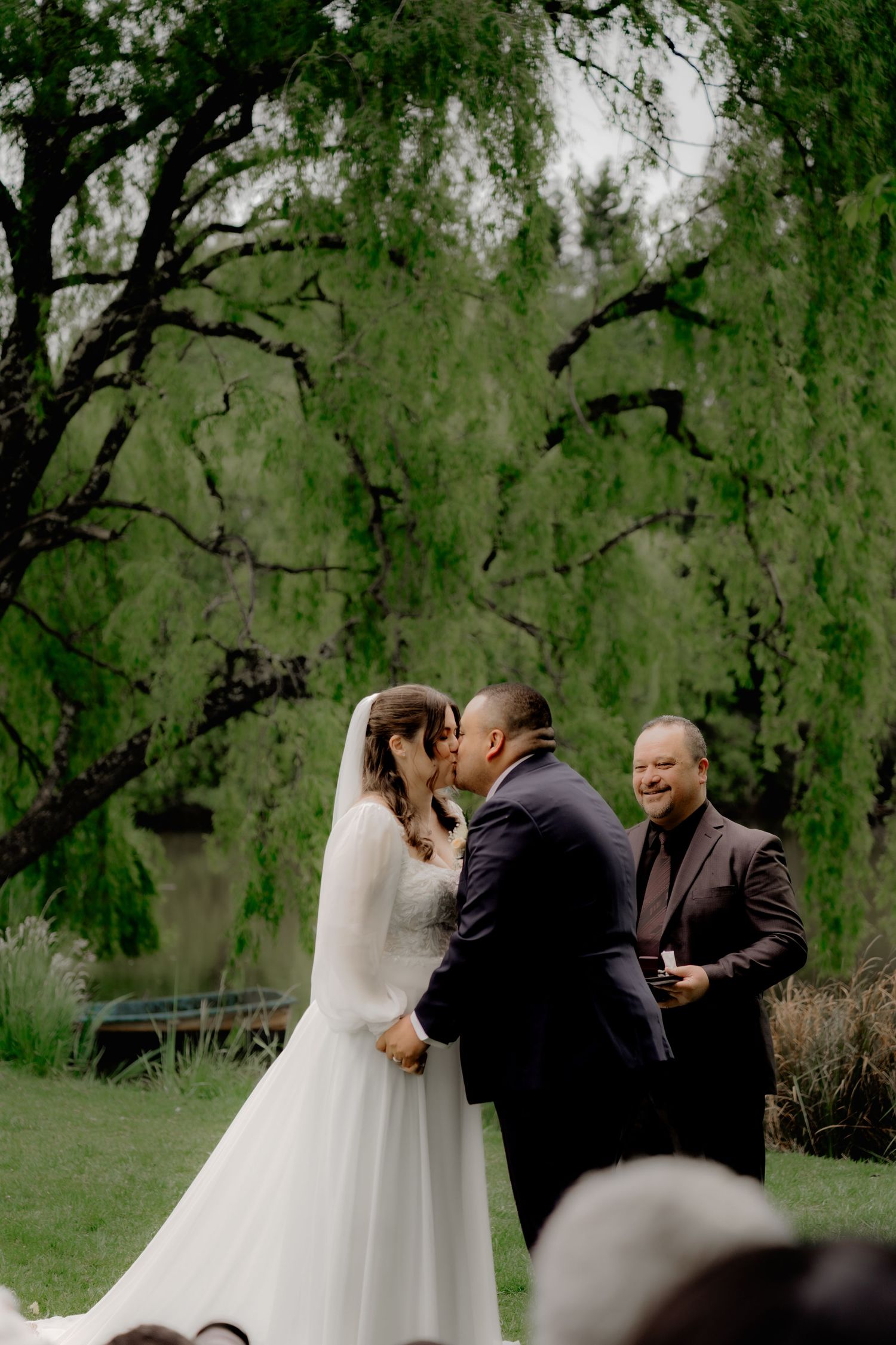 Bride and groom share first kiss at outdoor wedding ceremony under weeping willow tree with smiling officiant.