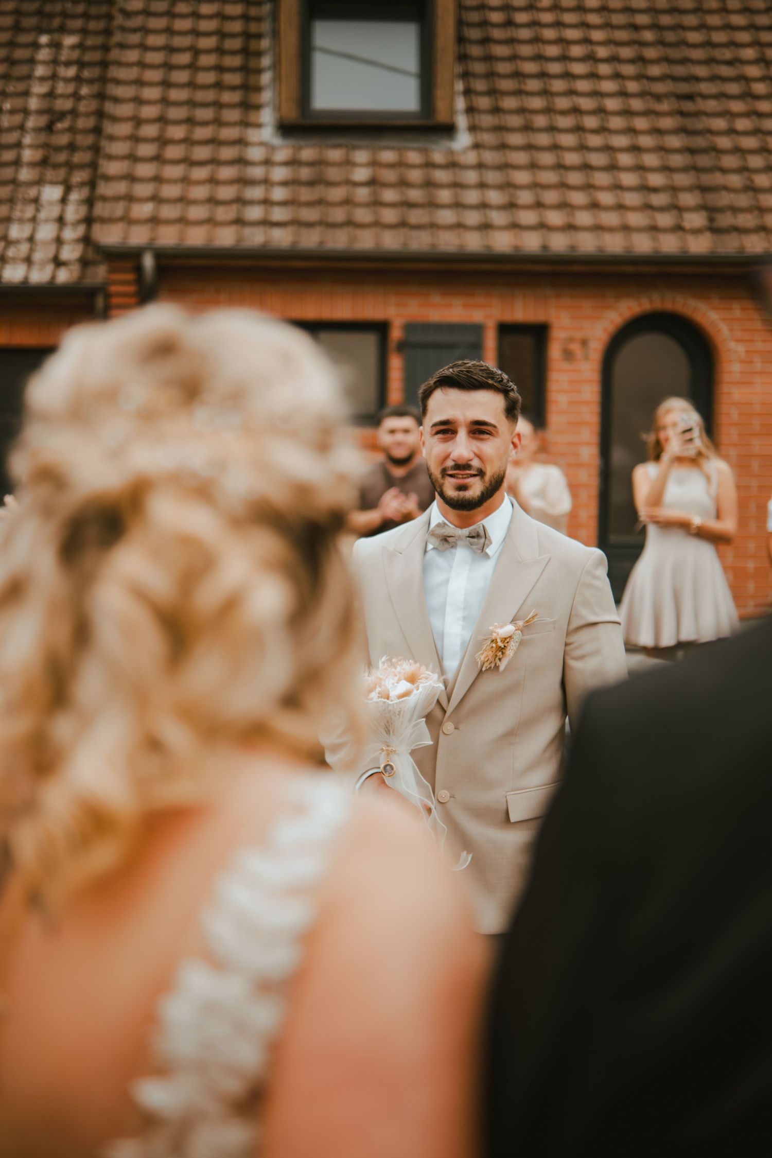 Emotional groom in beige suit with bow tie watches bride approach during outdoor wedding ceremony near brick building.