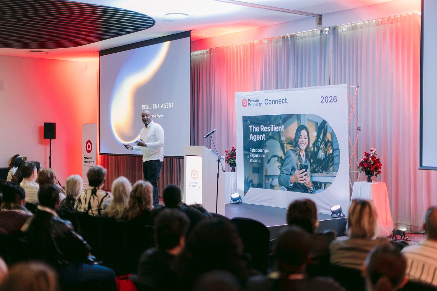 Speaker presenting at a corporate conference event in 2026 with audience and branded backdrop on stage.