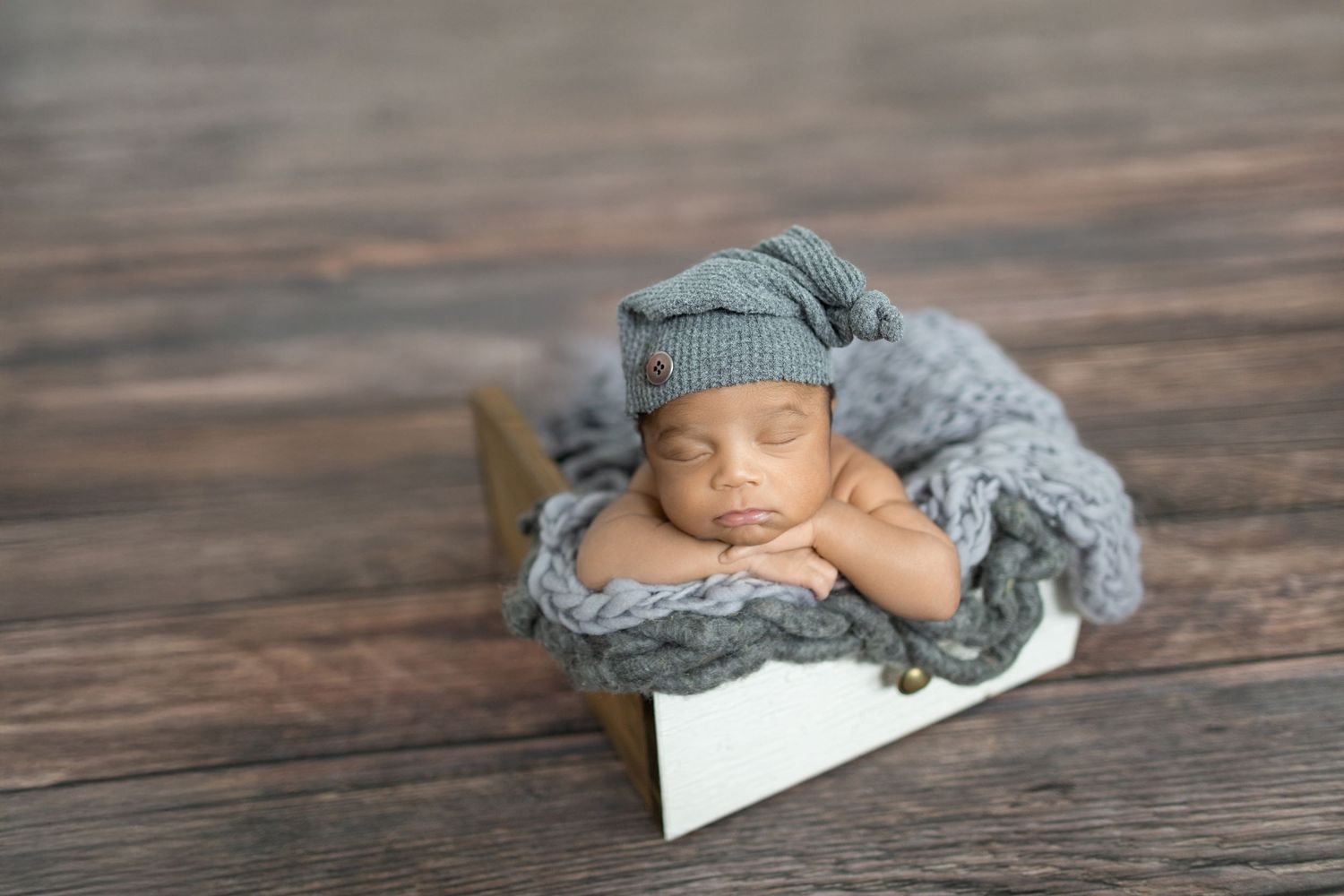 Sleeping newborn baby in gray knit hat resting in wooden box with soft gray wrap on rustic wood floor.