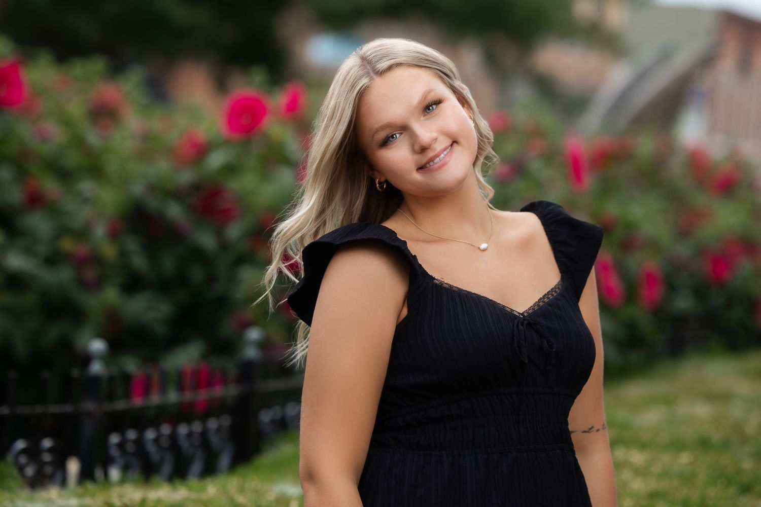 Young blonde woman in black mini dress posing outdoors near red roses and green hedges.