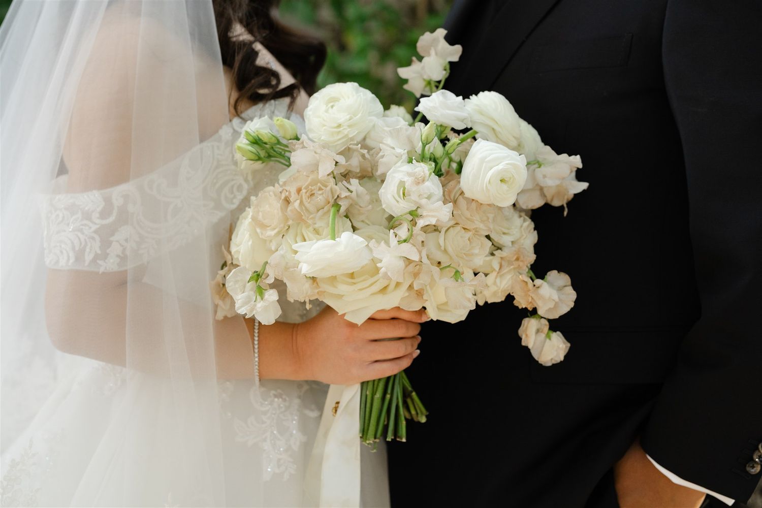 Bride and groom holding a lush white wedding bouquet with ranunculus and roses, bride wearing lace veil.