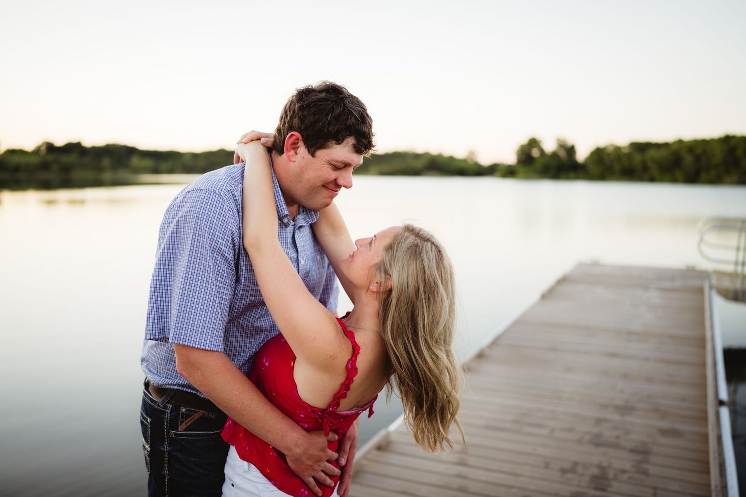 A couple holds hands and smiles at each other in a sunlit green meadow during a golden hour engagement photo session.