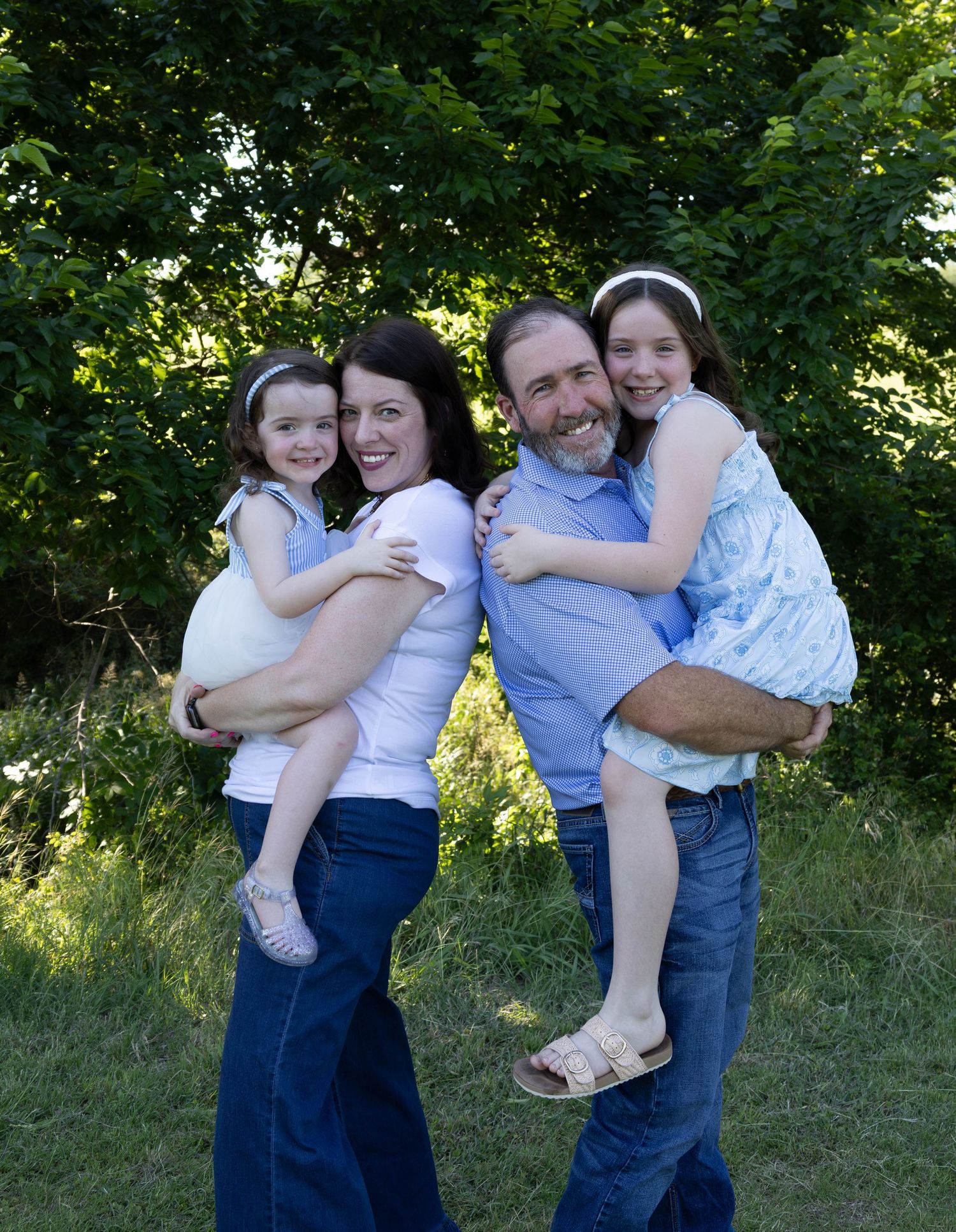 Family of four smiling outdoors on green grass, parents and two girls in white dresses, surrounded by lush trees.