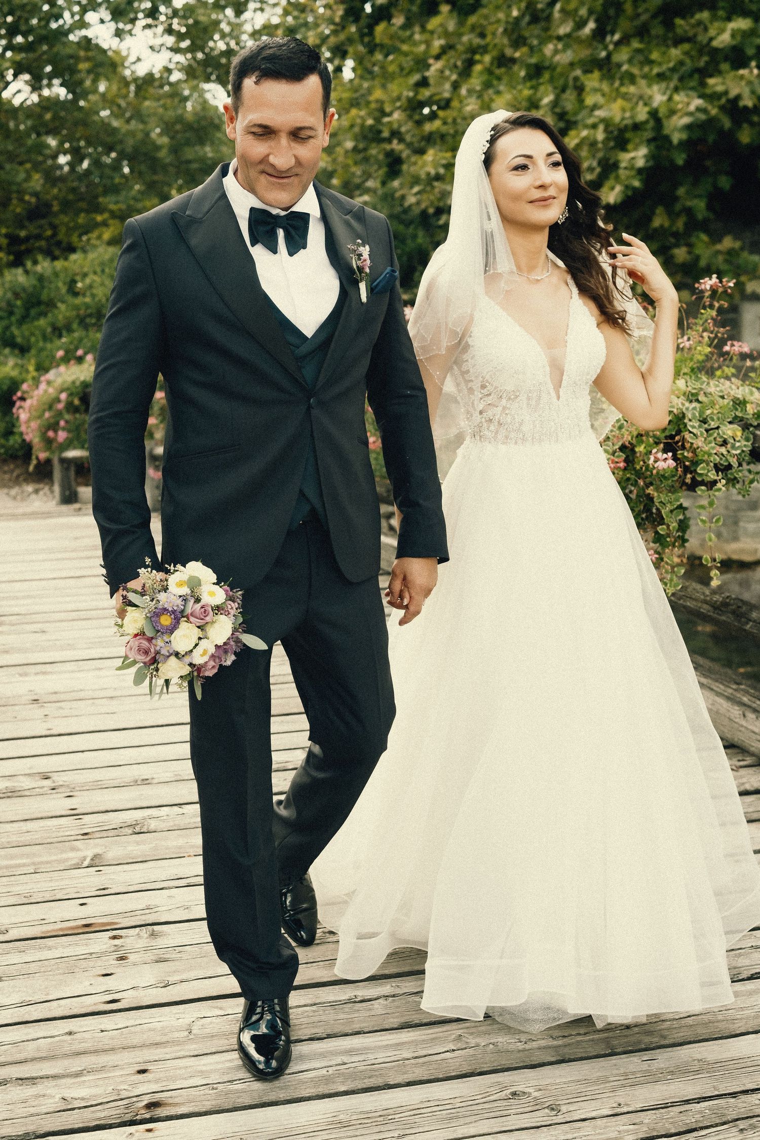 A bride and groom walk hand in hand across a wooden boardwalk in a garden setting.