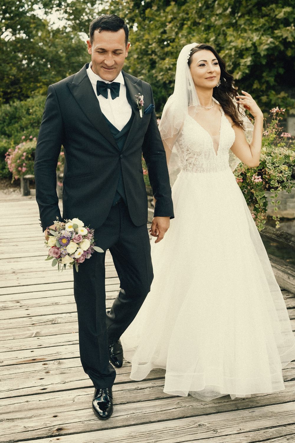 A bride and groom walk hand in hand across a wooden boardwalk in a garden setting.