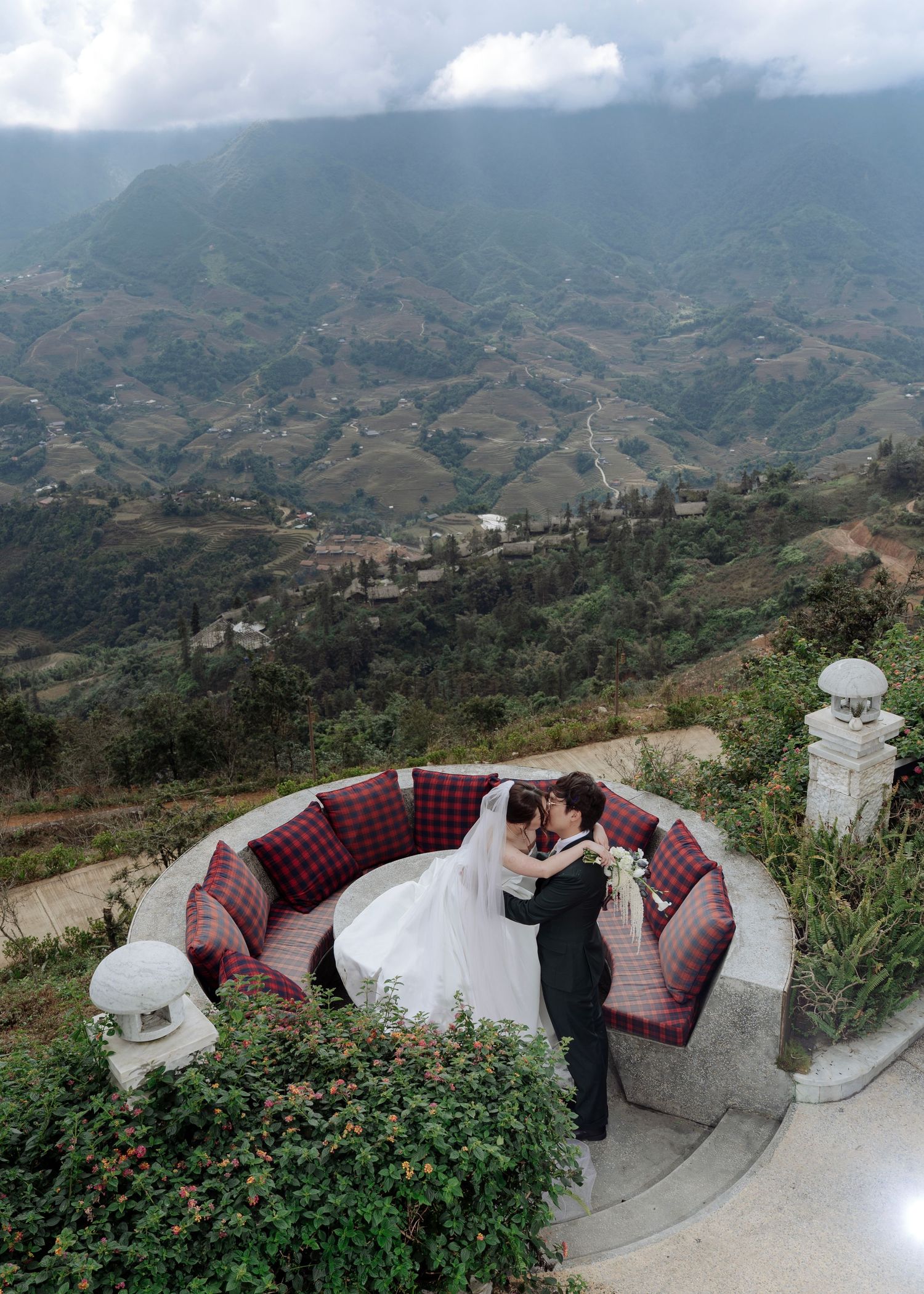 Bride and groom embrace on a circular red sofa terrace overlooking stunning mountain valley terraces in Sapa, Vietnam.