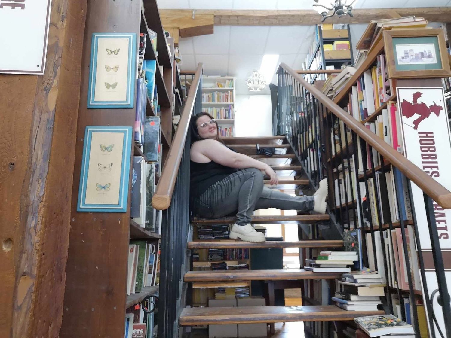 A person relaxes on wooden stairs in a cozy bookstore surrounded by shelves of books and framed butterfly artwork.