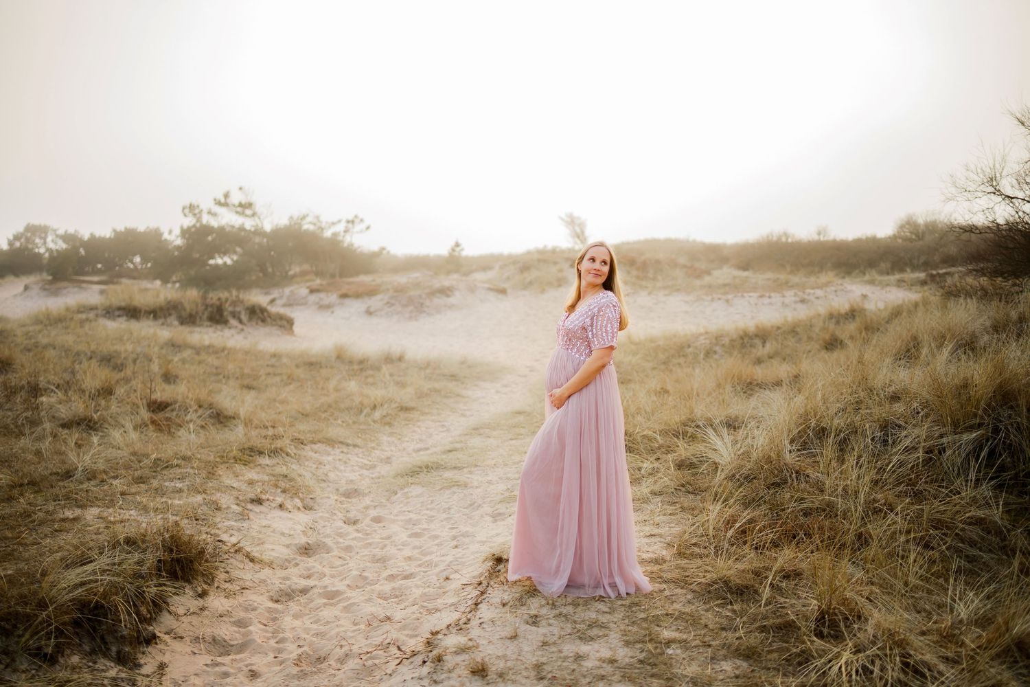Gravid kvinna i rosa flödande klänning poserar på sandig bana omgiven av gyllene gräs i mjukt solljus.