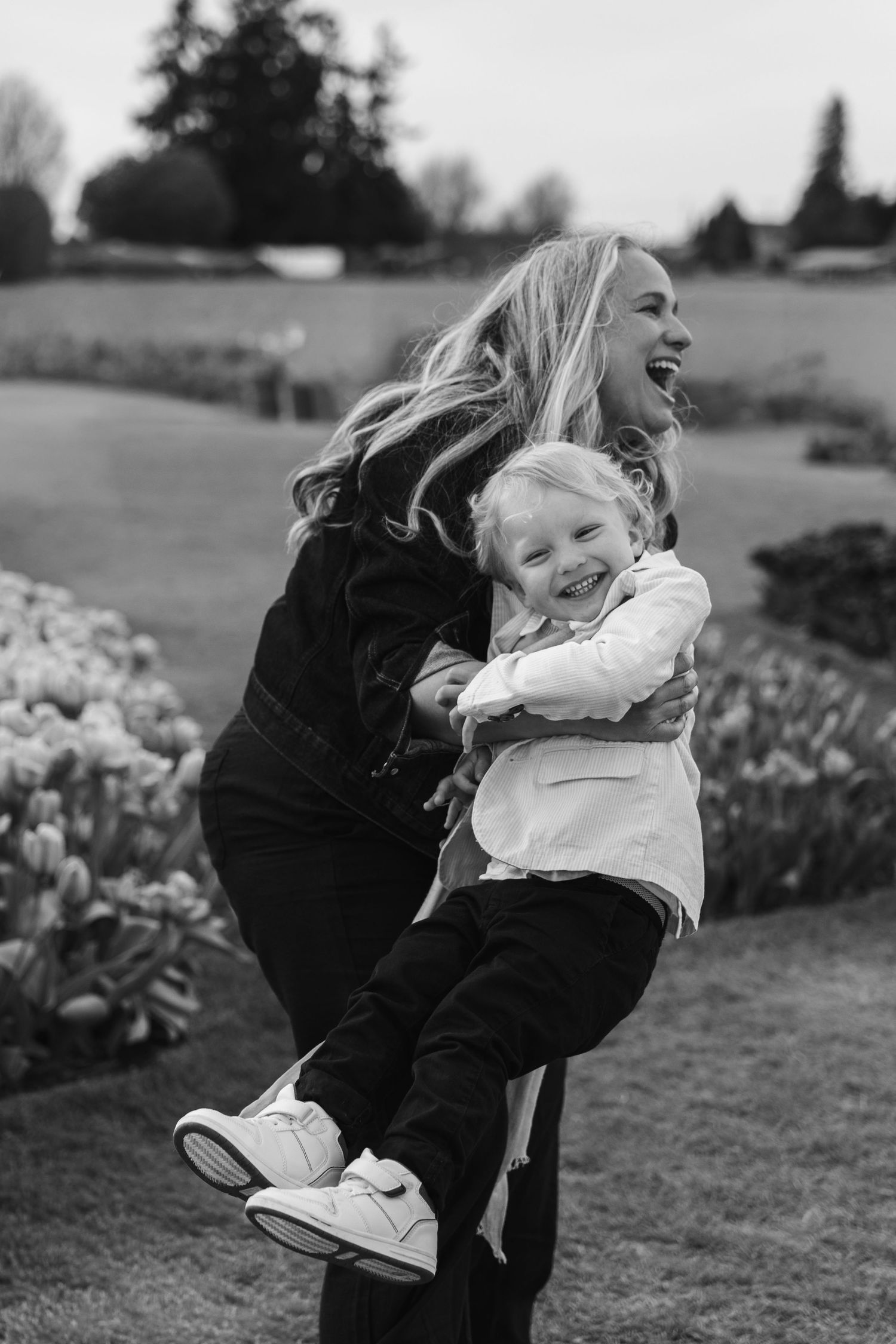 Black and white photo of a laughing woman lifting a happy toddler outdoors near a garden.
