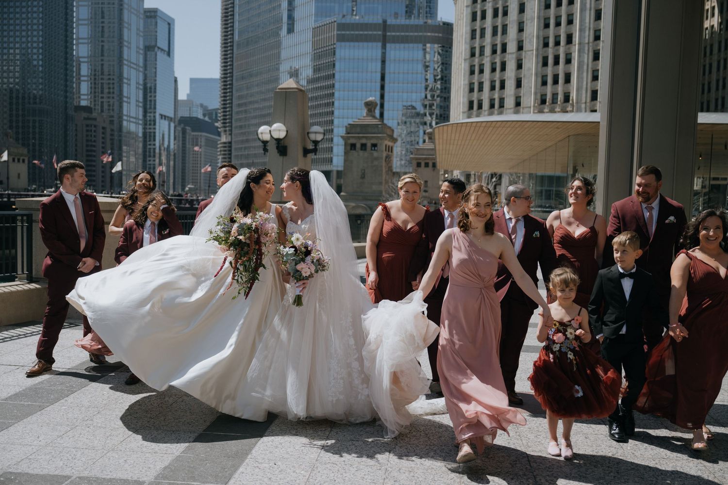 Two brides kiss while wedding party celebrates on a sunny Chicago riverwalk with city skyline backdrop.