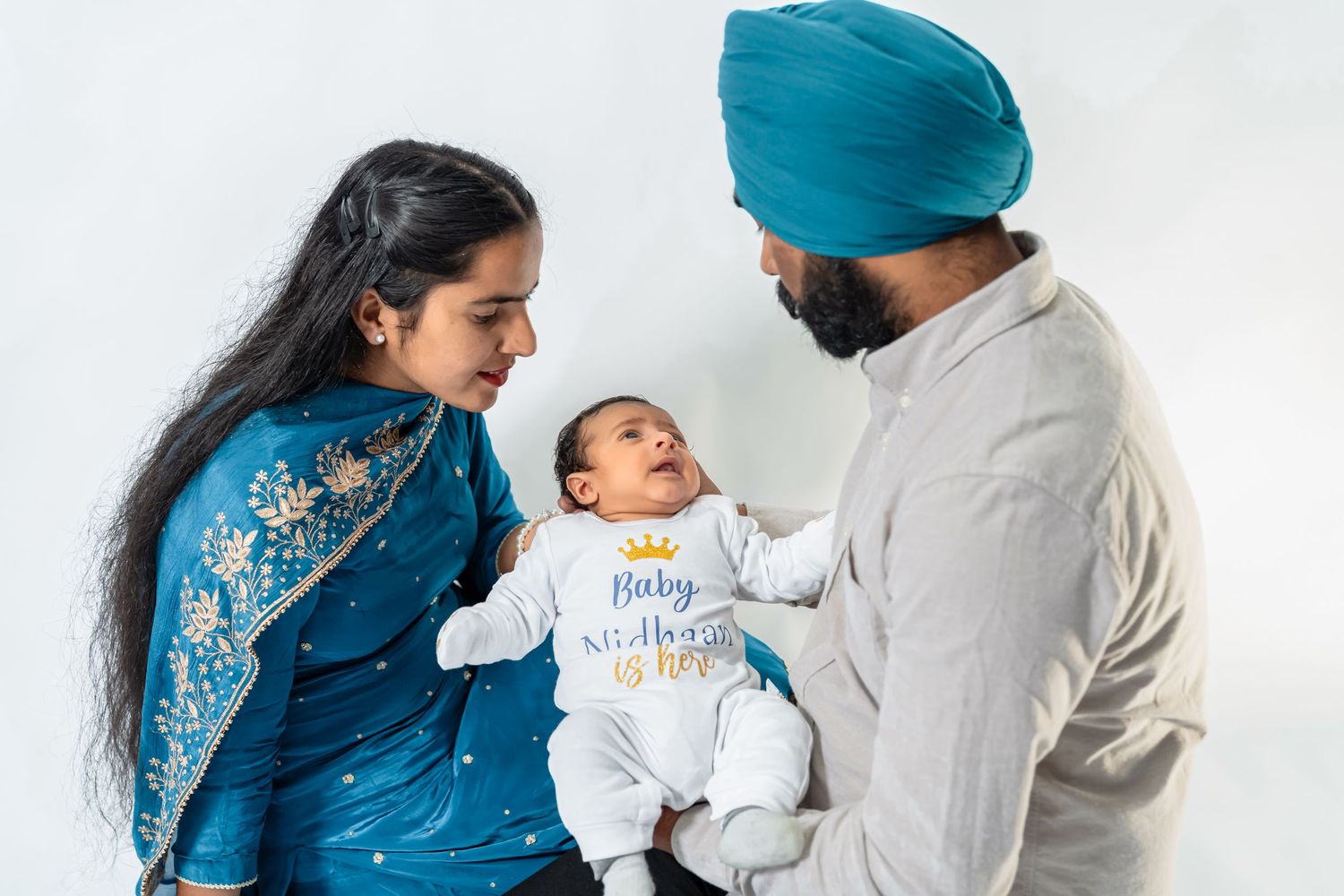 Sikh couple in traditional attire holding a newborn baby, posing together in front of a decorated backdrop.