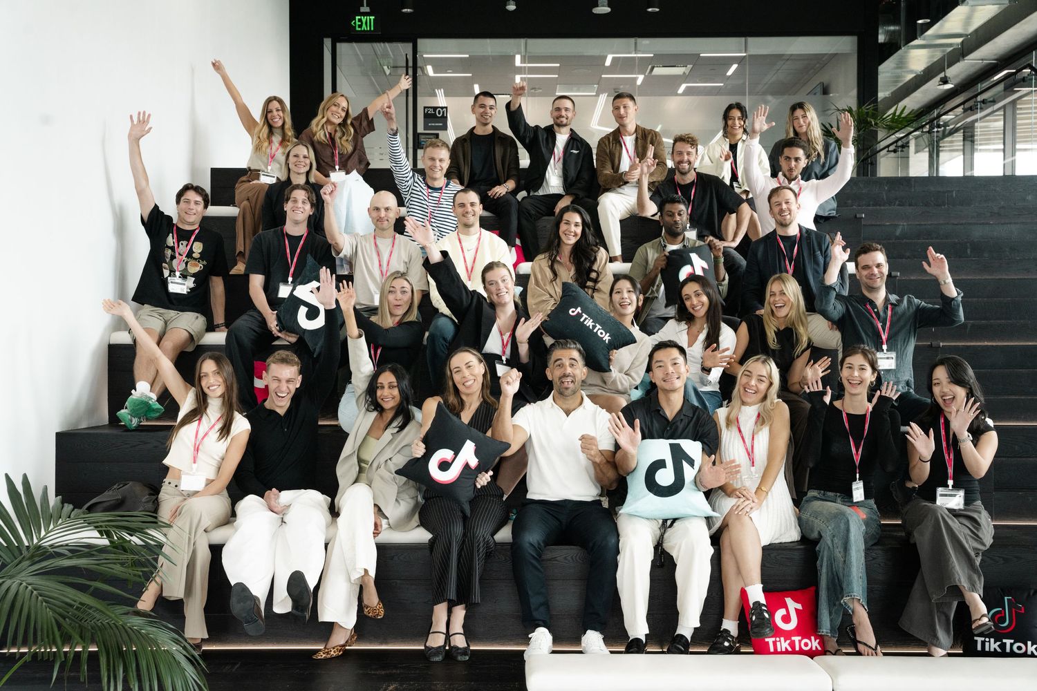 Large group of TikTok employees posing on stairs, smiling and waving, wearing branded black t-shirts in modern office.