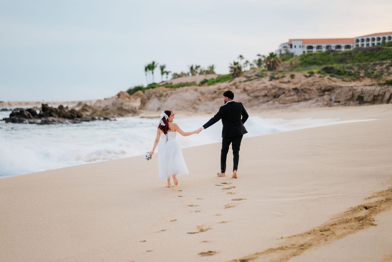 Bride and groom walk hand-in-hand along a sandy beach, with ocean waves and a resort in the background.