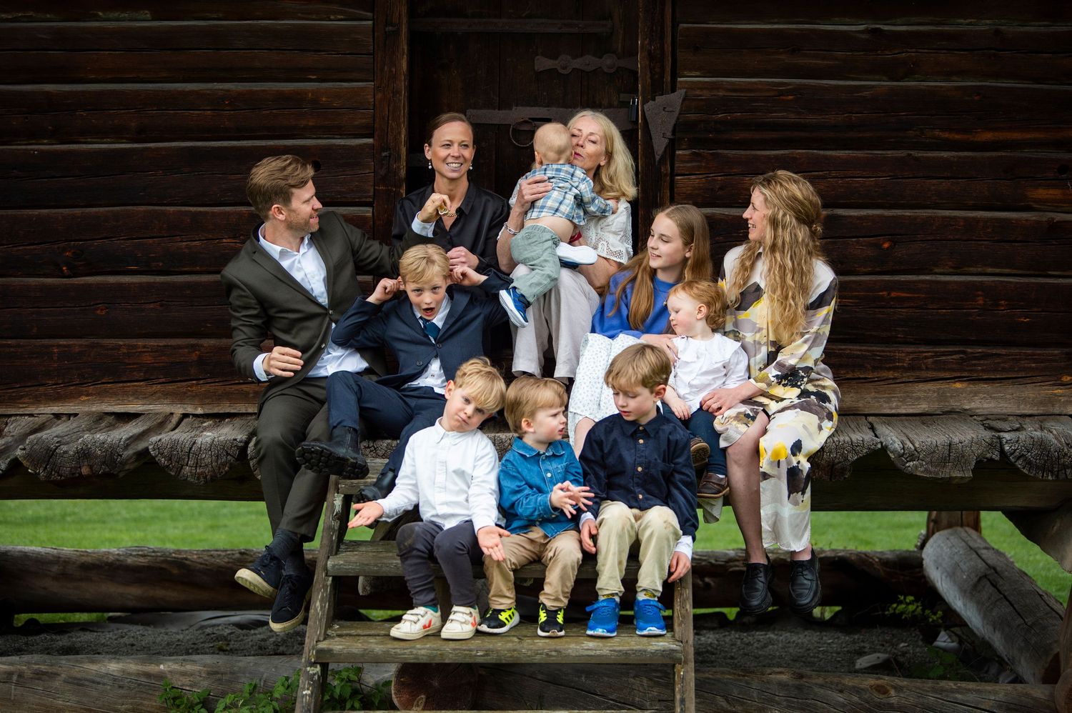 Large family group portrait with adults and children sitting on wooden steps outside a rustic log cabin.