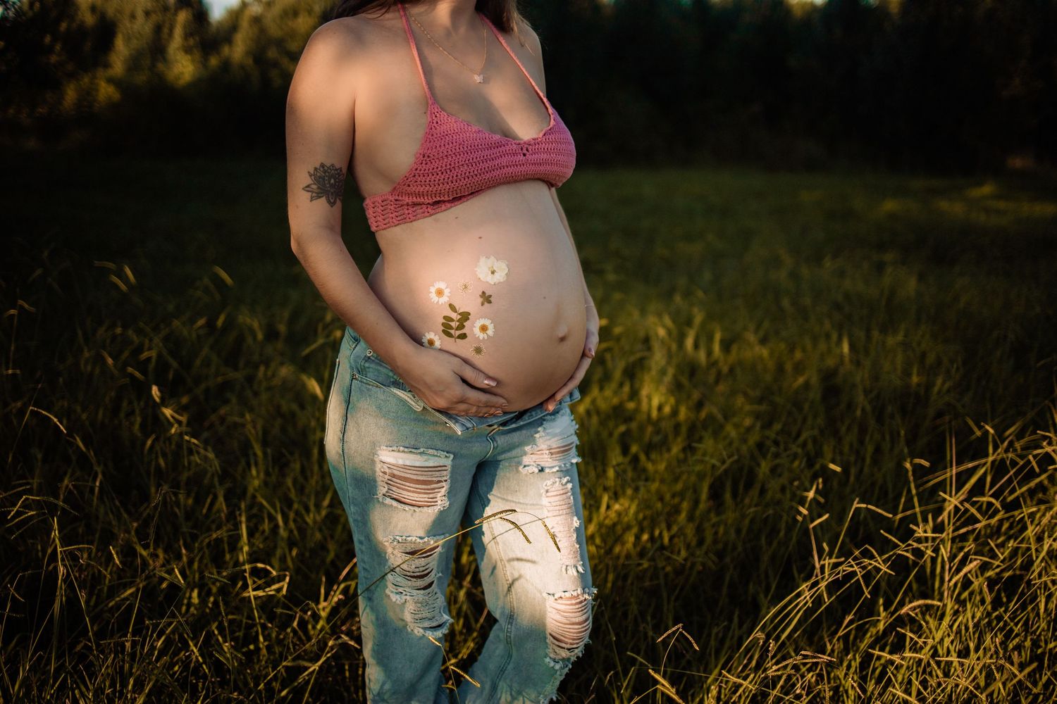 Pregnant woman in pink crop top and ripped jeans holding baby bump in sunlit field.