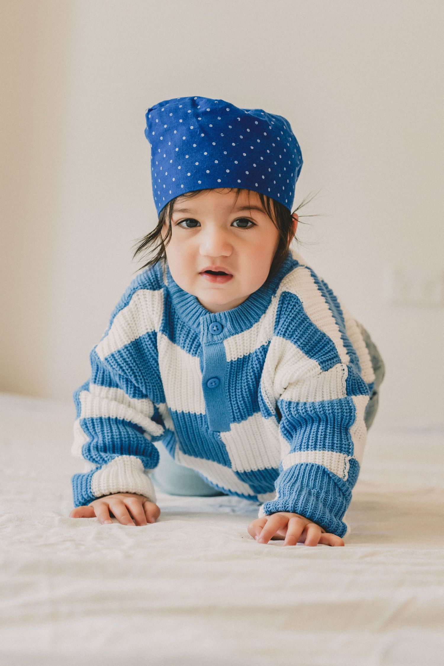 Cute baby in blue checkered knit sweater and polka dot hat crawling on white bed.