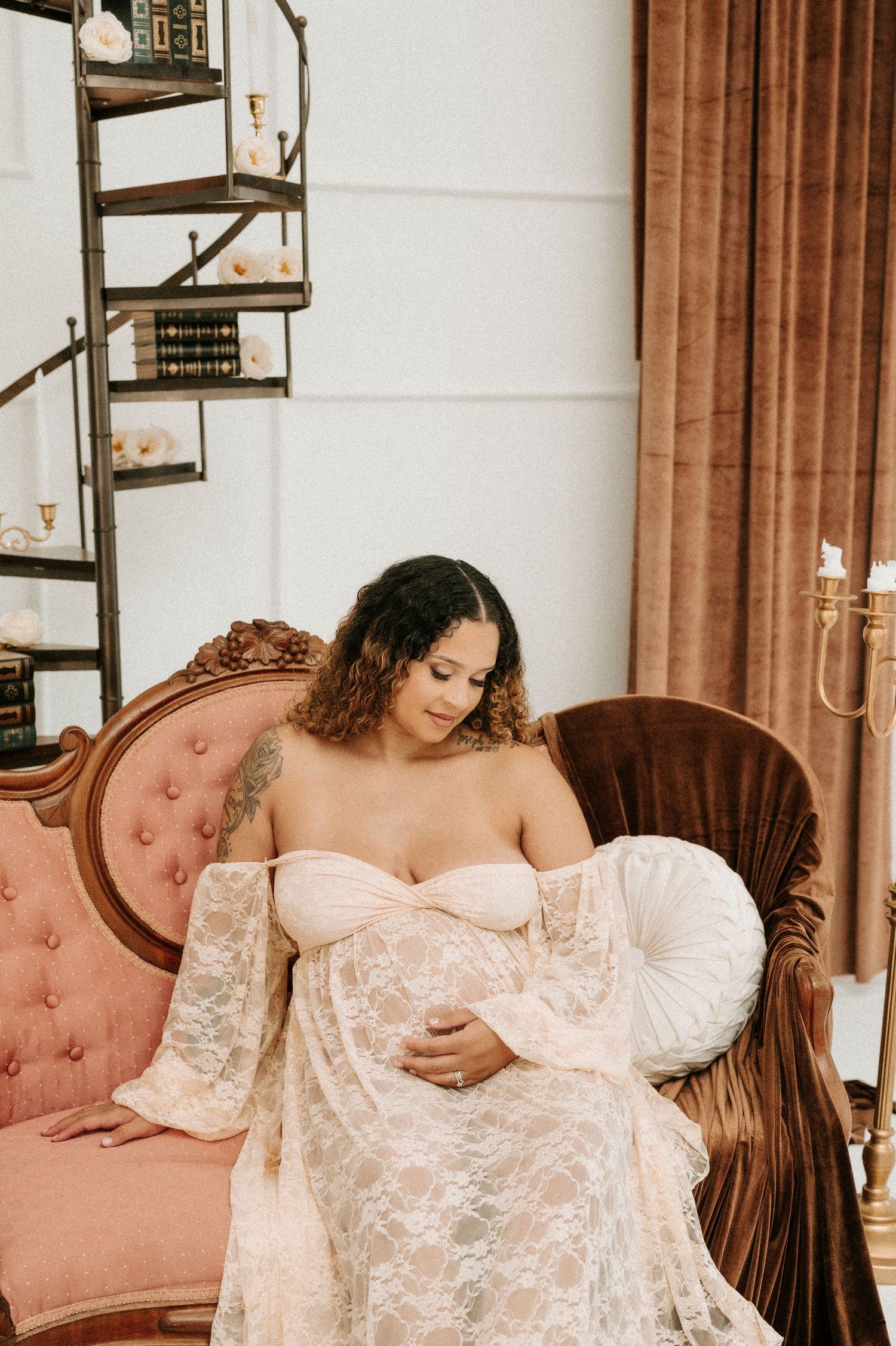 Pregnant woman in white lace off-shoulder gown seated on pink tufted sofa, cradling baby bump in elegant vintage-styled room.