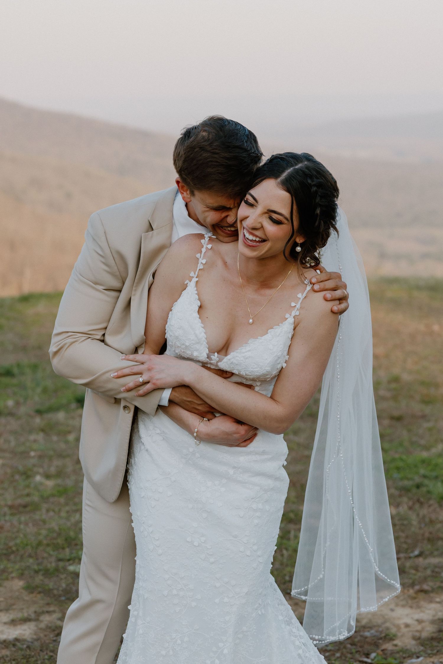 A romantic wedding couple embraces outdoors with mountains in the background during golden hour.