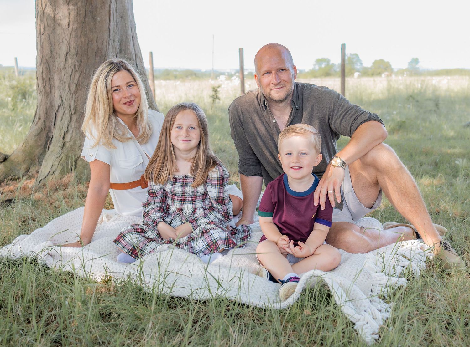 Family of four smiling together on a white blanket in a grassy field near a large tree on a sunny day.