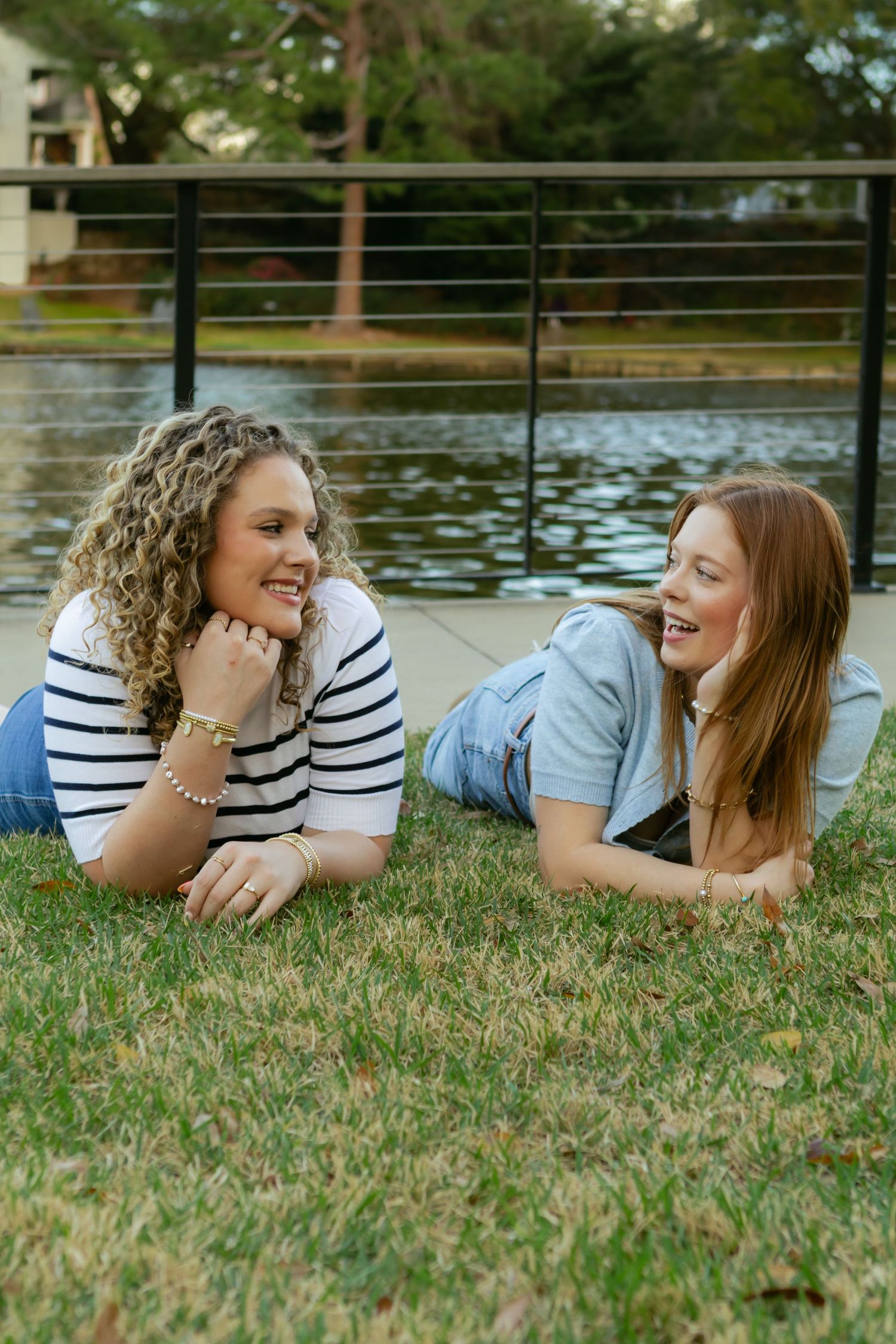 Two friends lying on grass by a lake share laughter and conversation on a sunny afternoon.