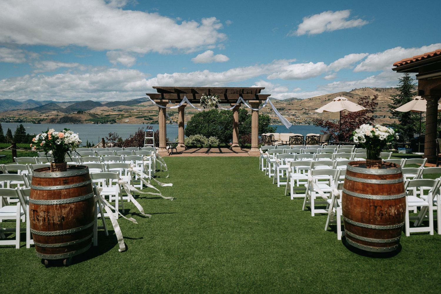 Outdoor winery wedding ceremony setup with white chairs, barrel arrangements, floral accents, and a wooden arch overlooking a scenic lake.