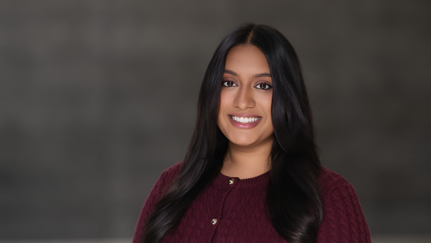 Professional headshot of a smiling woman in a burgundy sweater against a dark gray background.
