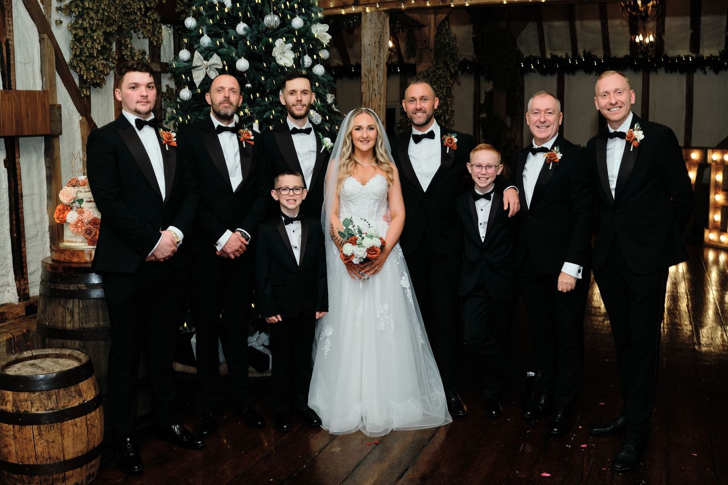 Bride and groom pose with groomsmen in black tuxedos at elegant winter wedding venue with festive decorations.
