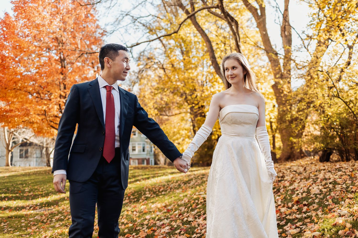 A couple in wedding attire holds hands walking through a park with vibrant autumn foliage in the background.