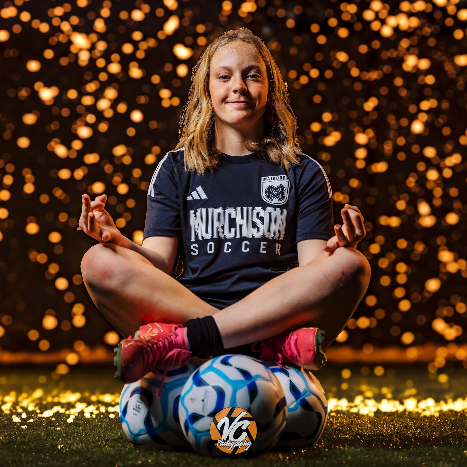 Young female soccer player sits cross-legged on soccer balls in Murchison Soccer jersey against golden bokeh backdrop.