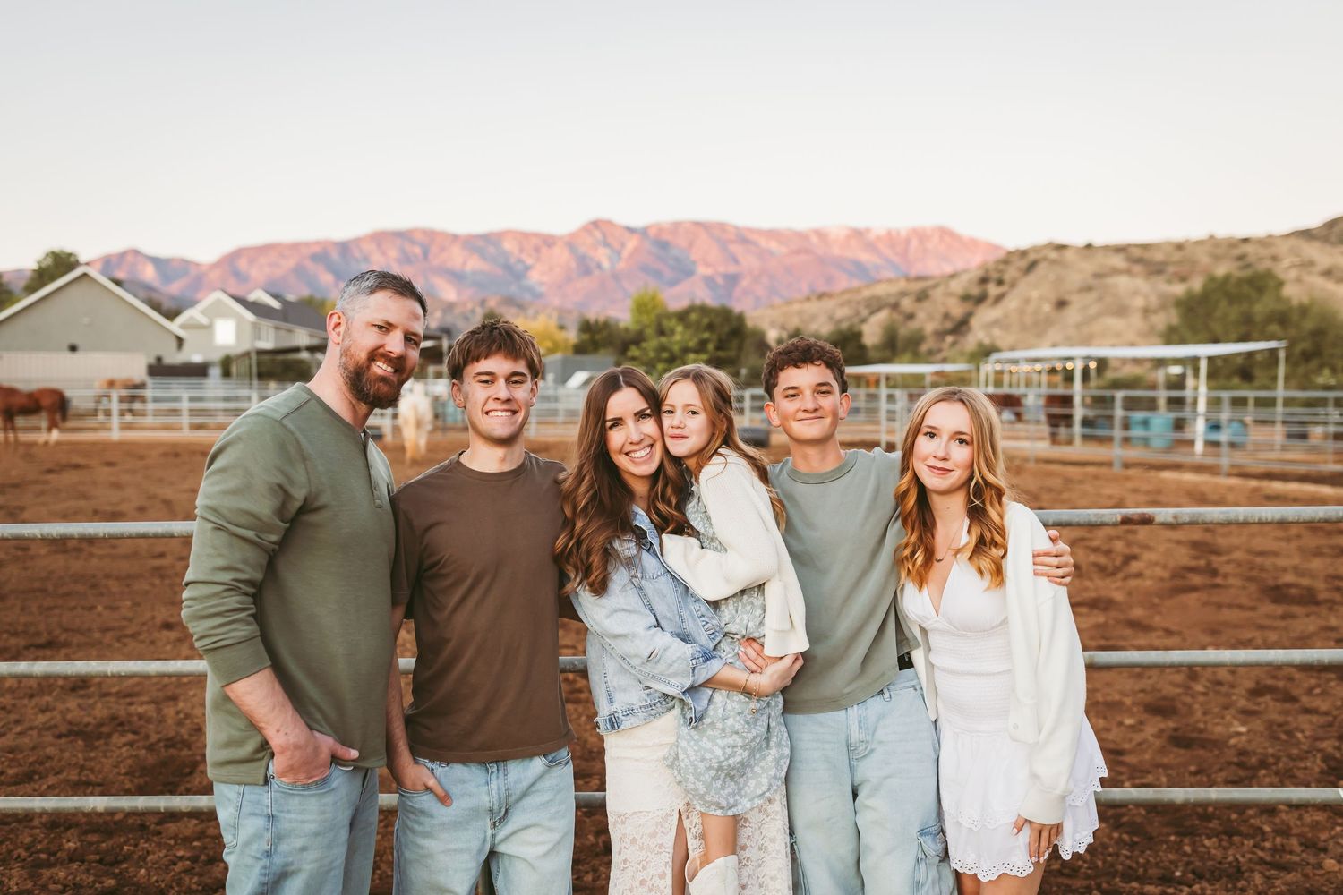 Family of six smiling together outdoors on a ranch with mountain views at sunset.